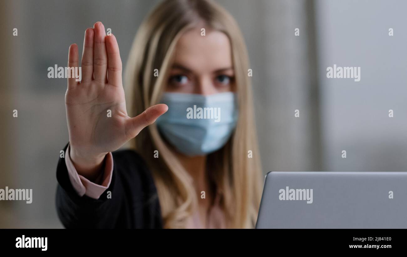 Ritratto in ufficio ragazza caucasica donna in maschera medica viso con laptop arrabbiato guardando la fotocamera mette la palma in avanti non stop guerra gesto proibizione Foto Stock