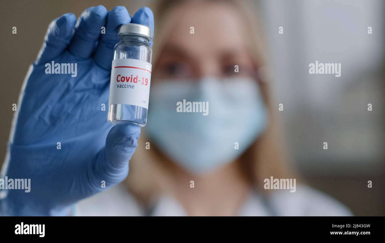 Primo piano donna specialista chimico biochimico donna medico infermiere scienziato in occhiali maschera medica e guanti in lattice guardando la fotocamera tenere bottiglia Foto Stock