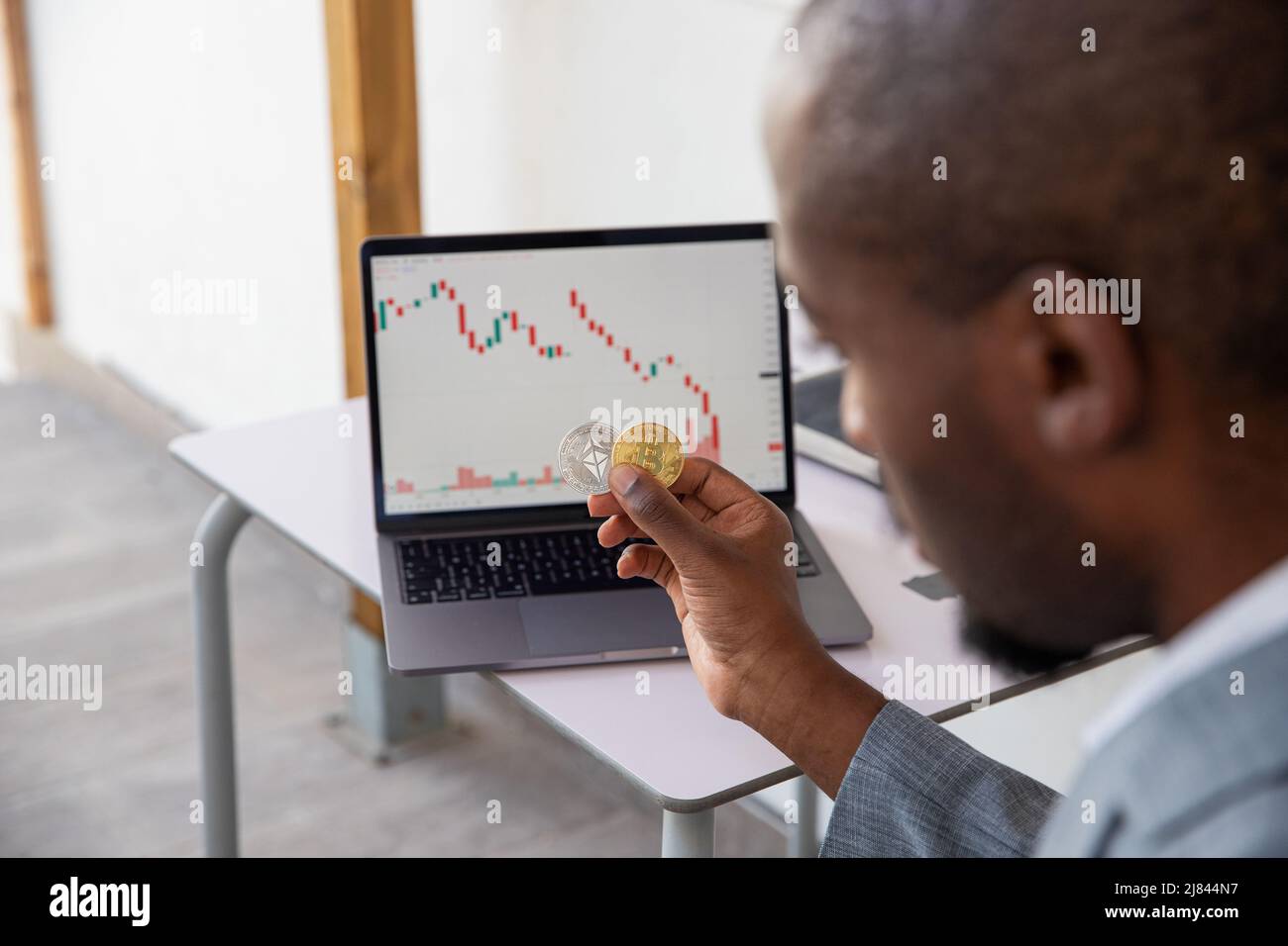 L'investitore di criptovaluta tiene in mano una moneta Bitcoin ed Ethereum, è triste perché il mercato sta cadendo e vede i grafici rossi sul suo lapto Foto Stock