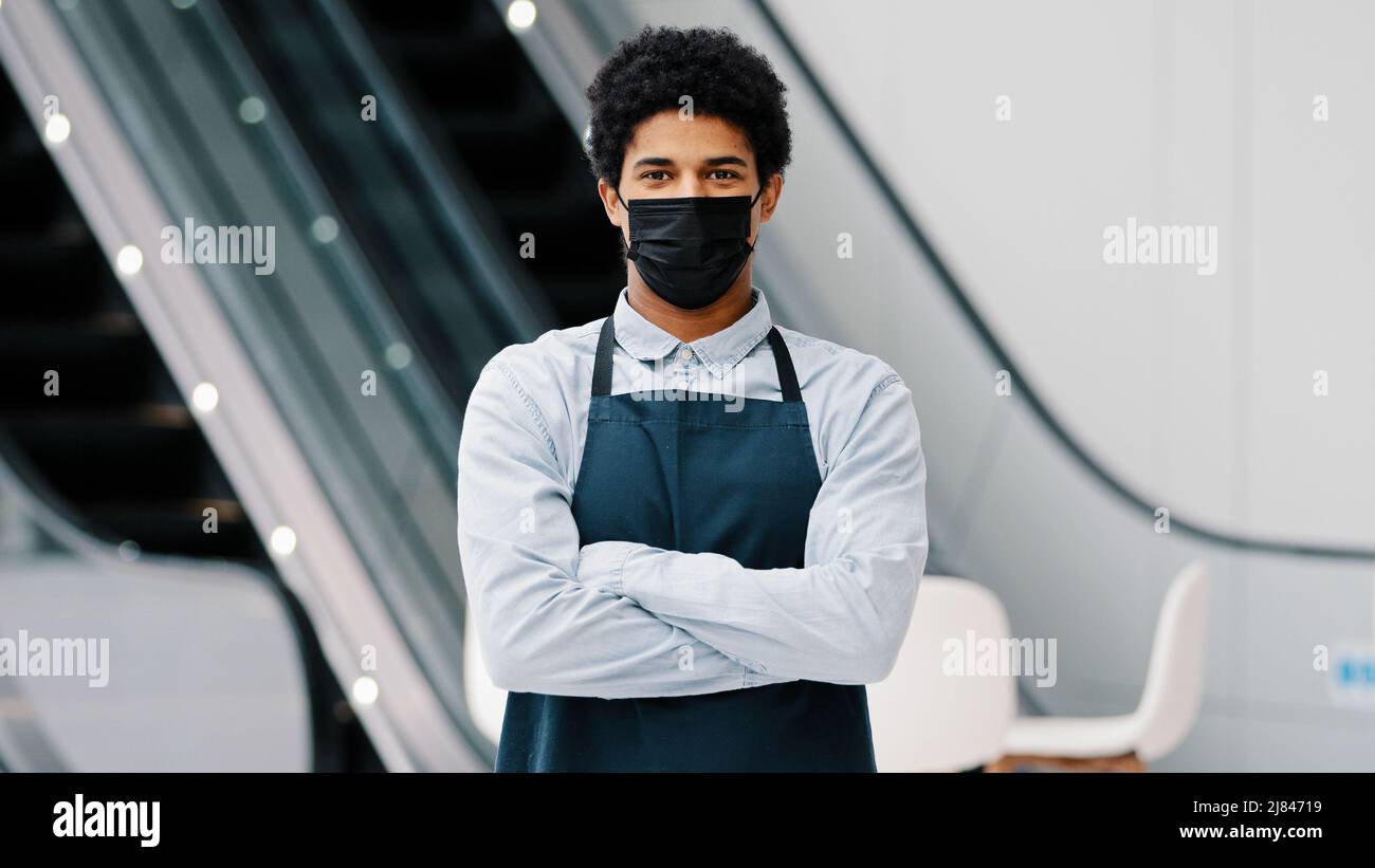 African american man guy ristorante cafe' uomo lavoratore in maschera medica che guarda la macchina fotografica che posa incrociando le braccia cameriere venditore indossare grembiule in piedi a. Foto Stock