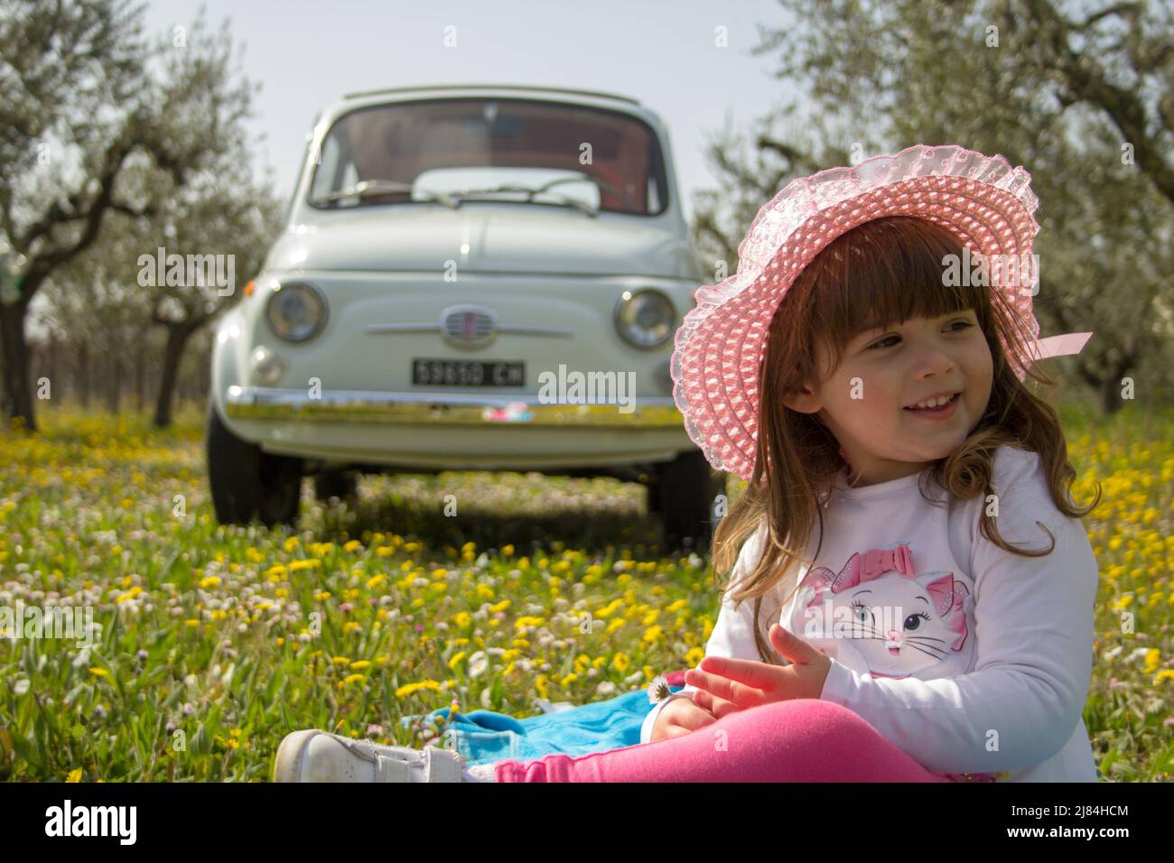 Foto di una sorridente bambina in un cappello seduto in un prato fiorito con un'auto d'epoca sullo sfondo durante un viaggio in Italia Foto Stock
