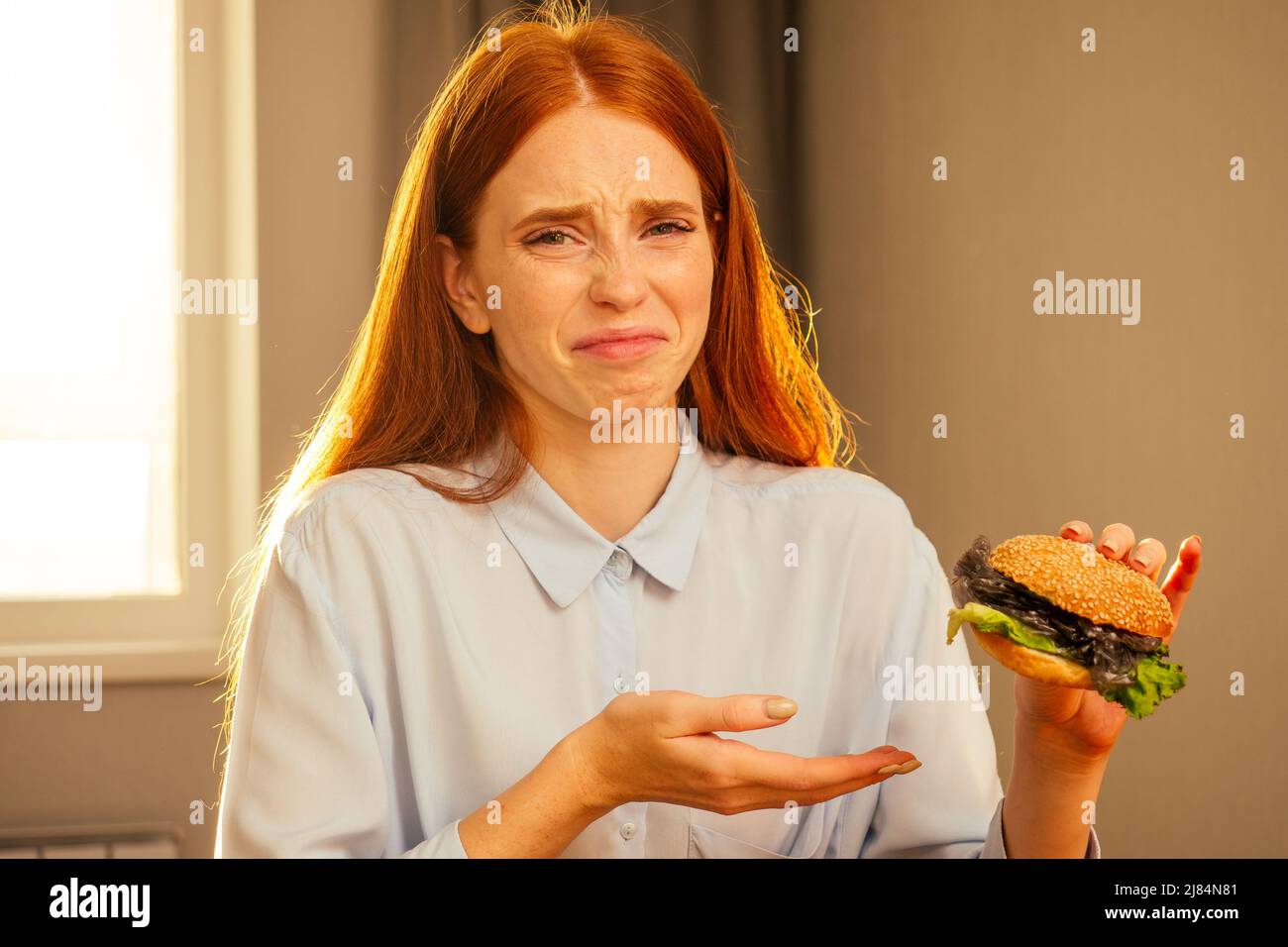 ragazza zenzero rossa che mangia hamburger finto con sesamo in panna e pellicola di cellophane di plastica spazzatura, acqua potabile da tazza monouso con tubo non-eco a. Foto Stock