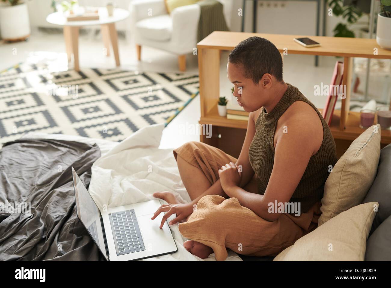 Giovane donna nera focalizzata con taglio buzz seduta sul letto e utilizzando il touchpad laptop in studio flat Foto Stock