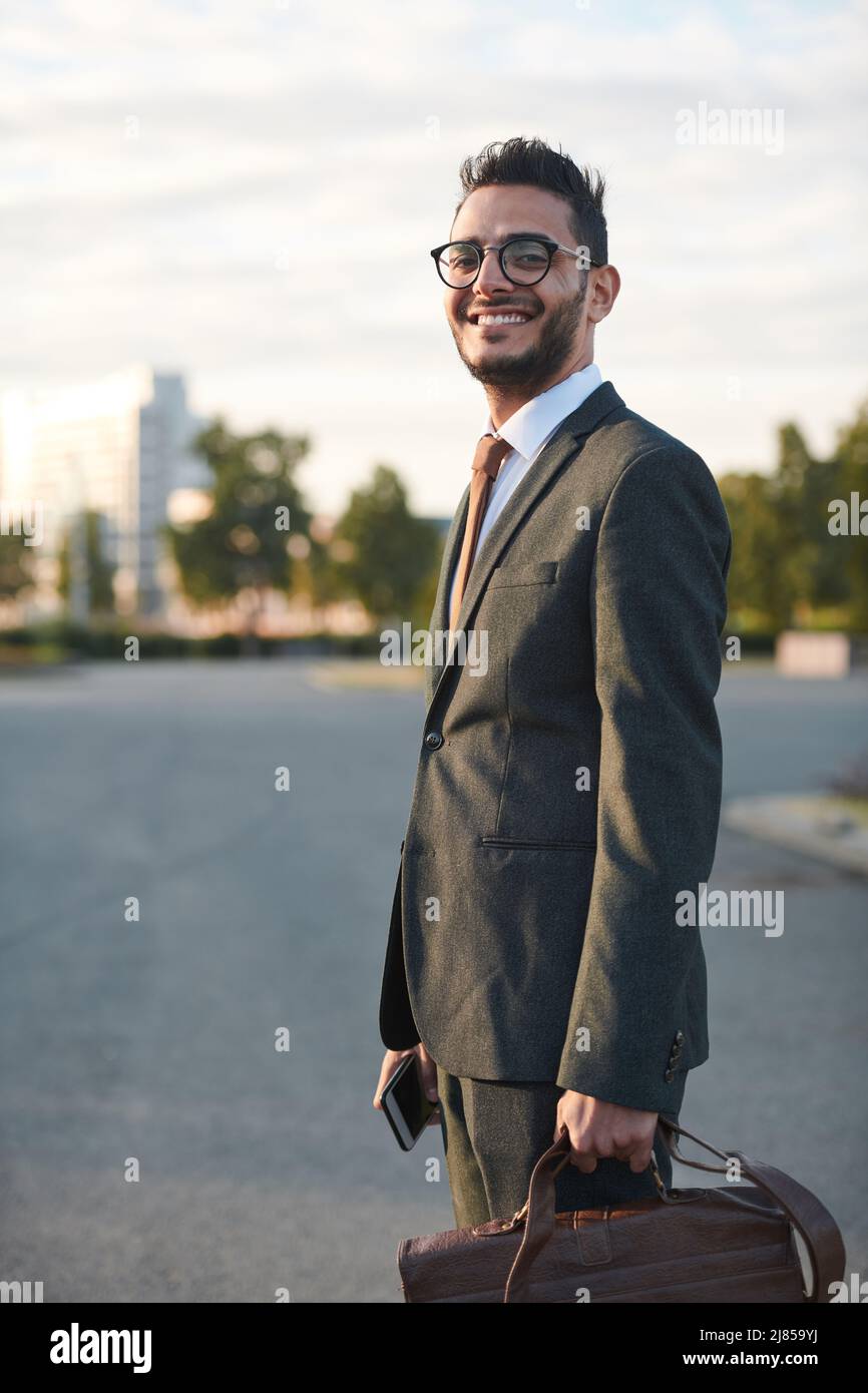 Ritratto di positivo giovane uomo d'affari arabo con barba che porta la valigetta mentre si va al lavoro Foto Stock