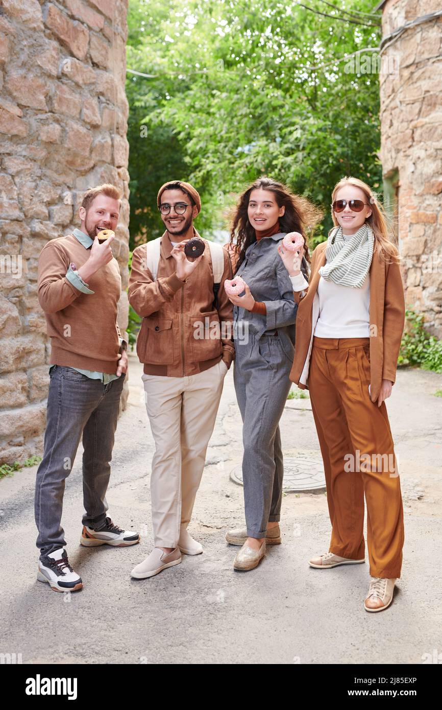 Gruppo di giovani amici multietnici positivi che mangiano gustose ciambelle mentre camminano sulla città vecchia Foto Stock