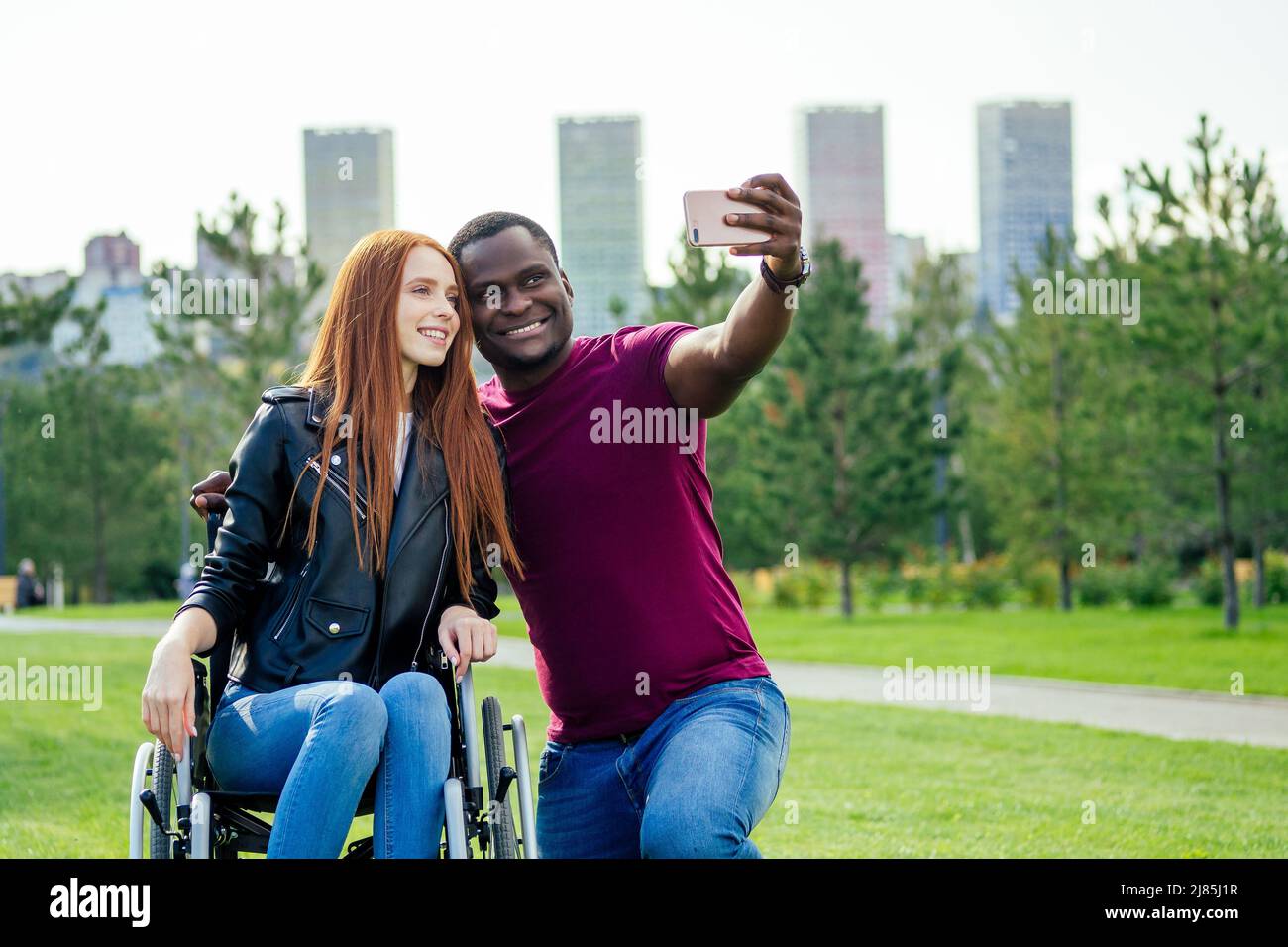 afro uomo americano seduto su sedia a rotelle, la sua ragazza di zenzero redhaired che rotola passeggino nel parco autunnale. avendo data romantica.making un selfie su Foto Stock