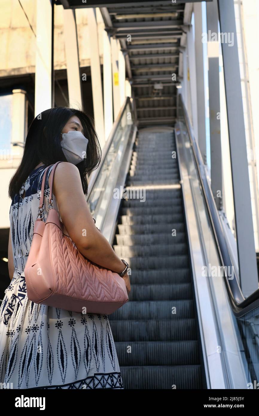 La vista posteriore di una donna asiatica con una maschera facciale bianca, indossando un abito bianco e blu sta salendo su una scala mobile per una stazione del treno sopraelevato al mattino. Foto Stock