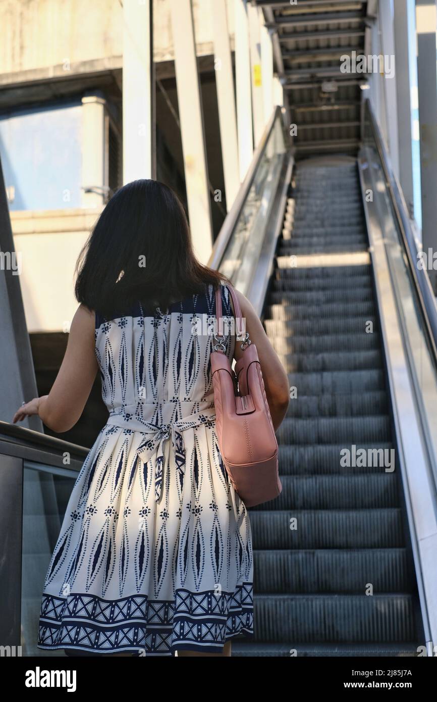 La vista posteriore di una donna asiatica in abito bianco e blu sta salendo su una scala mobile per una stazione del treno sopraelevato al mattino. Foto Stock