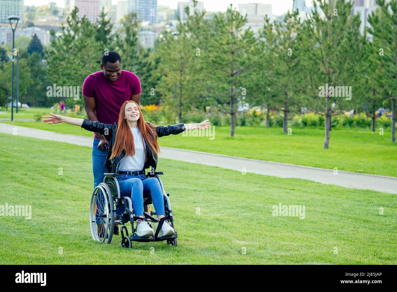 afro american uomo seduto su sedia a rotelle, la sua ragazza zenzero rossa girello rolling nel parco autunnale. avendo data romantica Foto Stock