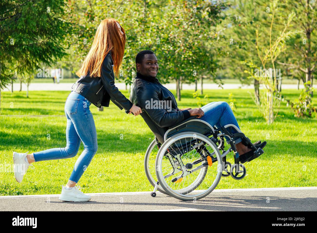 afro american uomo seduto su sedia a rotelle, la sua ragazza zenzero rossa girello rolling nel parco autunnale. avendo data romantica Foto Stock