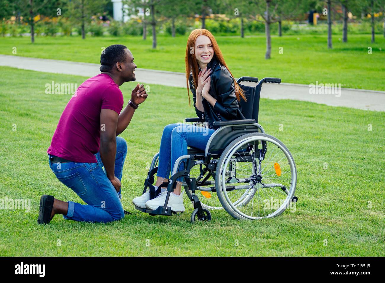afro american uomo matrimonio proposta dando un anello al suo rosso zenzero girlfriend.she seduta su sedia ruota e sorpreso e stupito Foto Stock