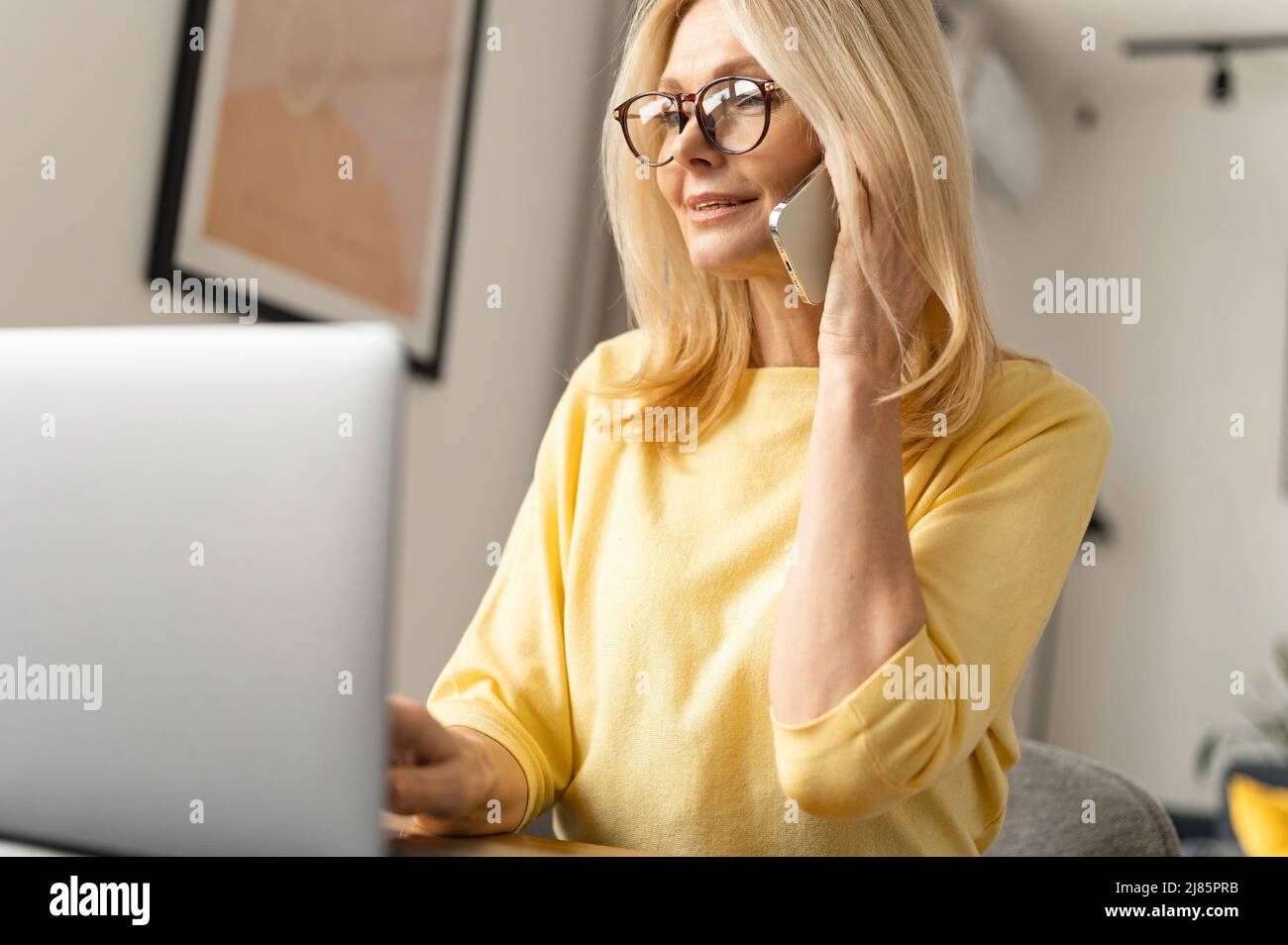 Sorridente donna anziana lavora da casa. Parla al telefono mentre si siede al tavolo davanti al portatile e al notebook Foto Stock