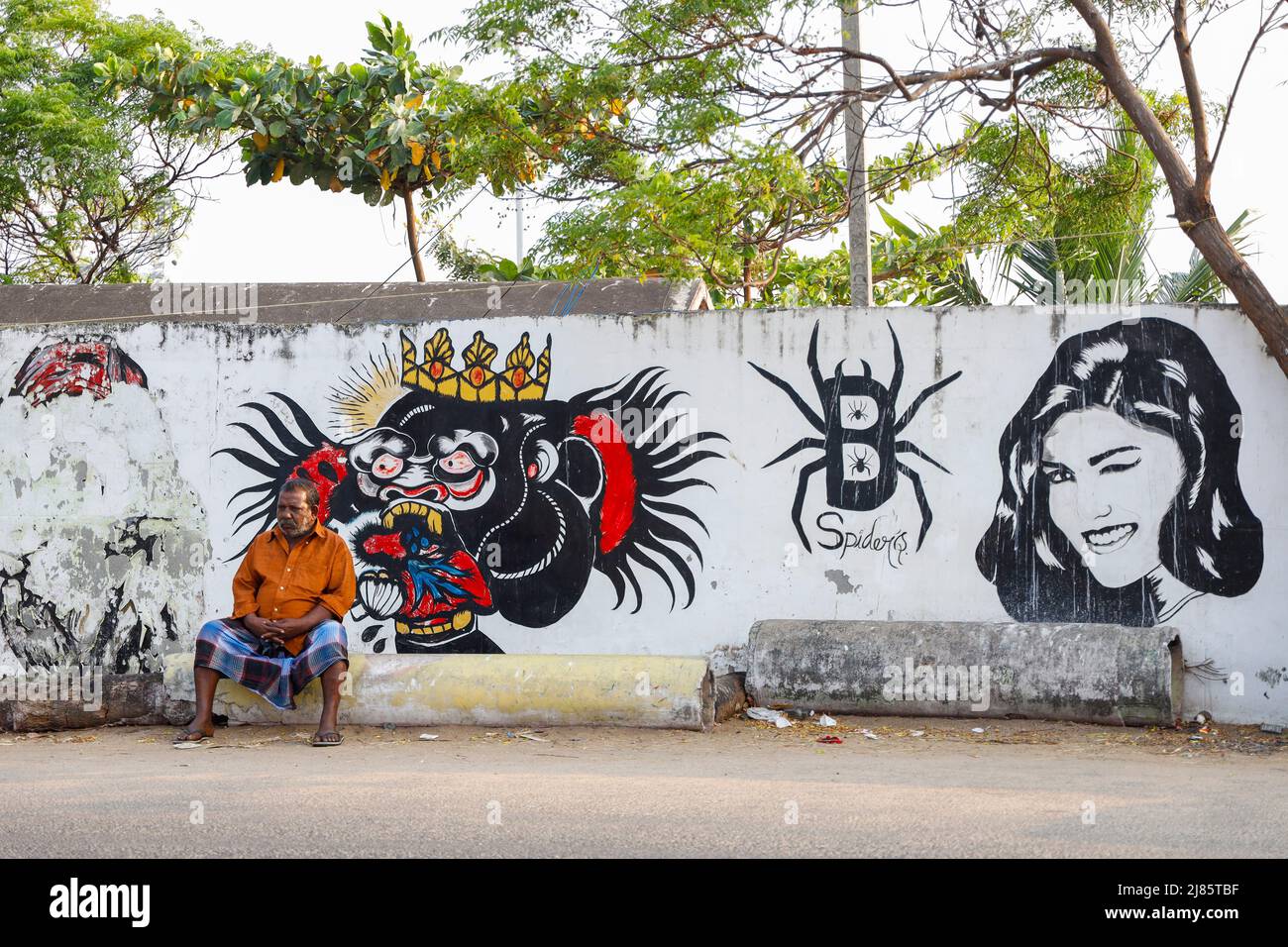 Vecchio uomo seduto di fronte ad un muro coperto di graffiti a Tangassery, Thangassery, Kerala, India. Foto Stock