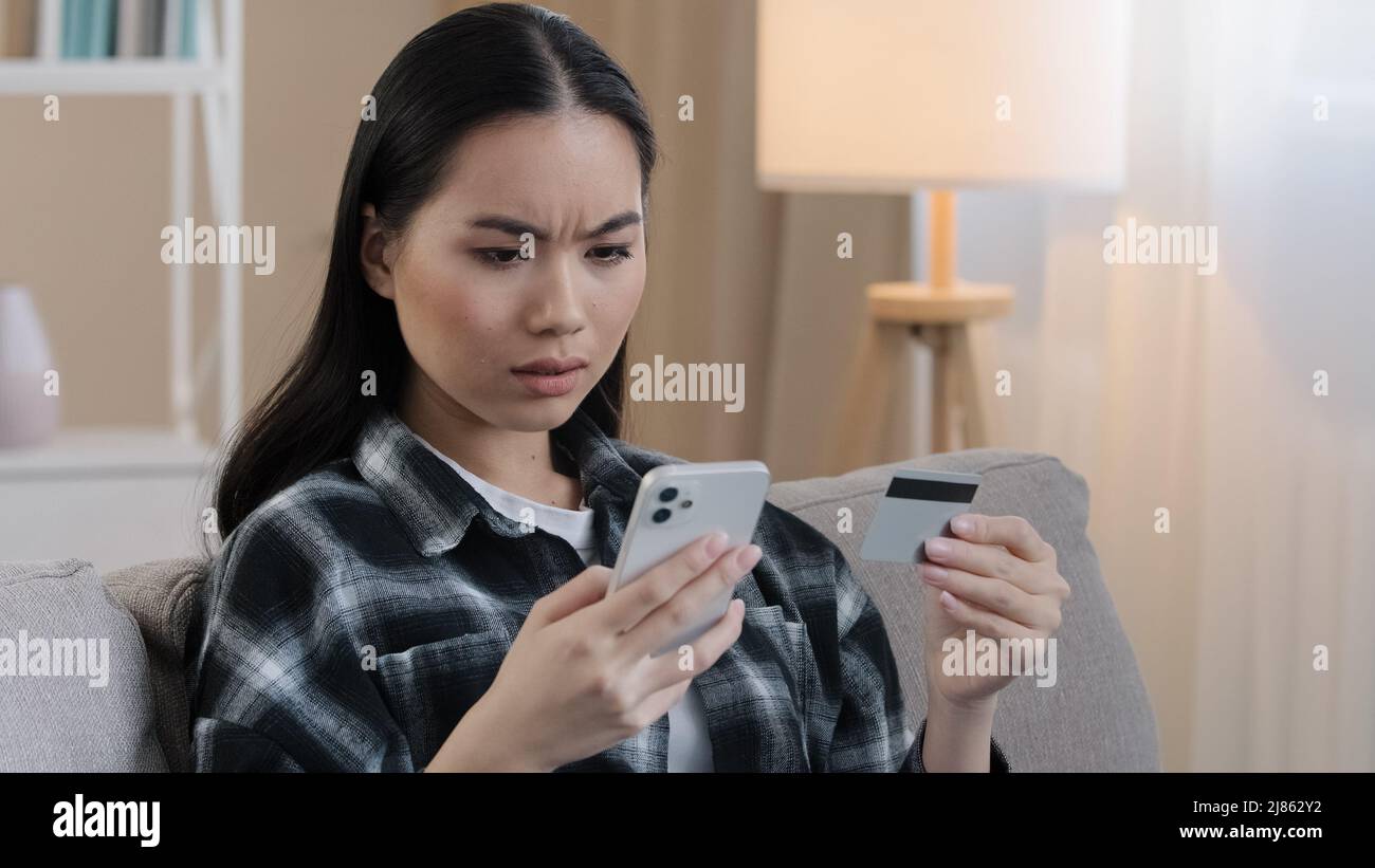 Donna asiatica arrabbiata seduta sul divano a casa tenere la carta di credito utilizzare il telefono cellulare provare a pagare con e-bank app problemi di pagamento errore bancario errore di denaro Foto Stock