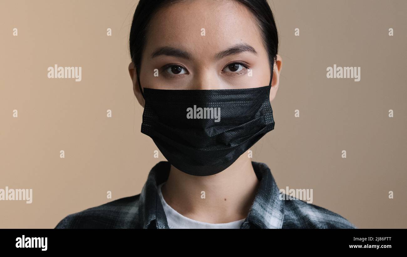 Ritratto interno chiuso viso espressione femmina viso asiatico razza donna male seria ragazza in nero maschera medica protettiva guardando fotocamera pandemic Foto Stock