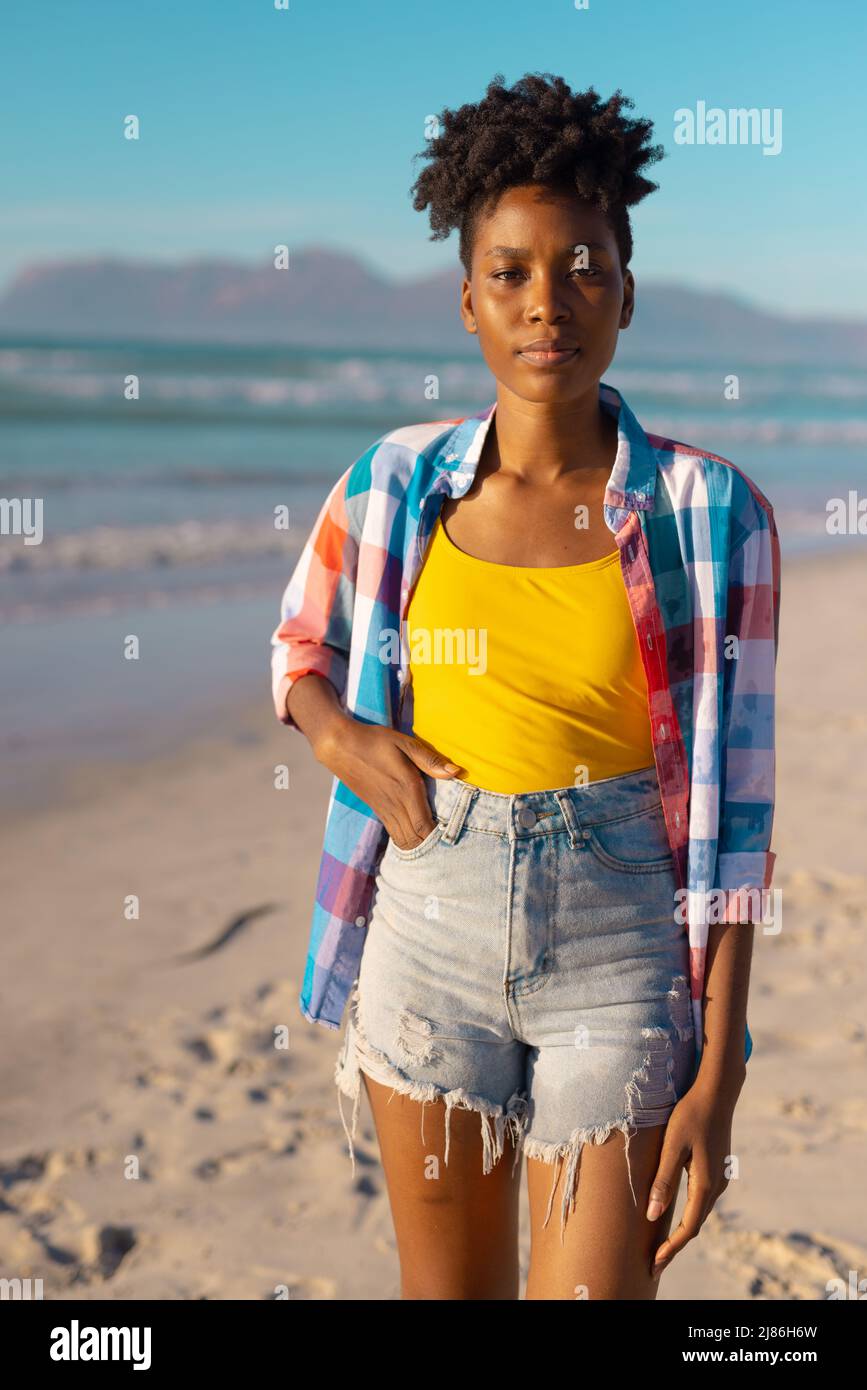 Ritratto di giovane afroamericana con capelli corti e mano in tasca contro il mare e il cielo blu Foto Stock