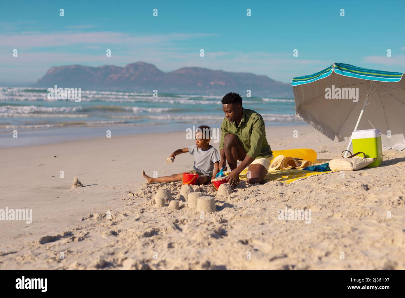 Giovane africano americano che fa castelli di sabbia con figlio in spiaggia contro il mare e il cielo in giorno di sole Foto Stock