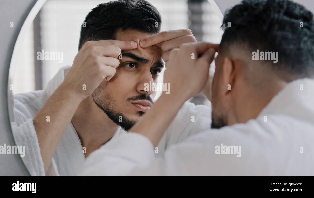 Infelice arabo indiano arabo indiano uomo bearded guardando nello specchio della stanza da bagno sensibilità preoccupata circa il problema della pelle facciale acne. Ragazzo ansioso millennial che stringe Foto Stock