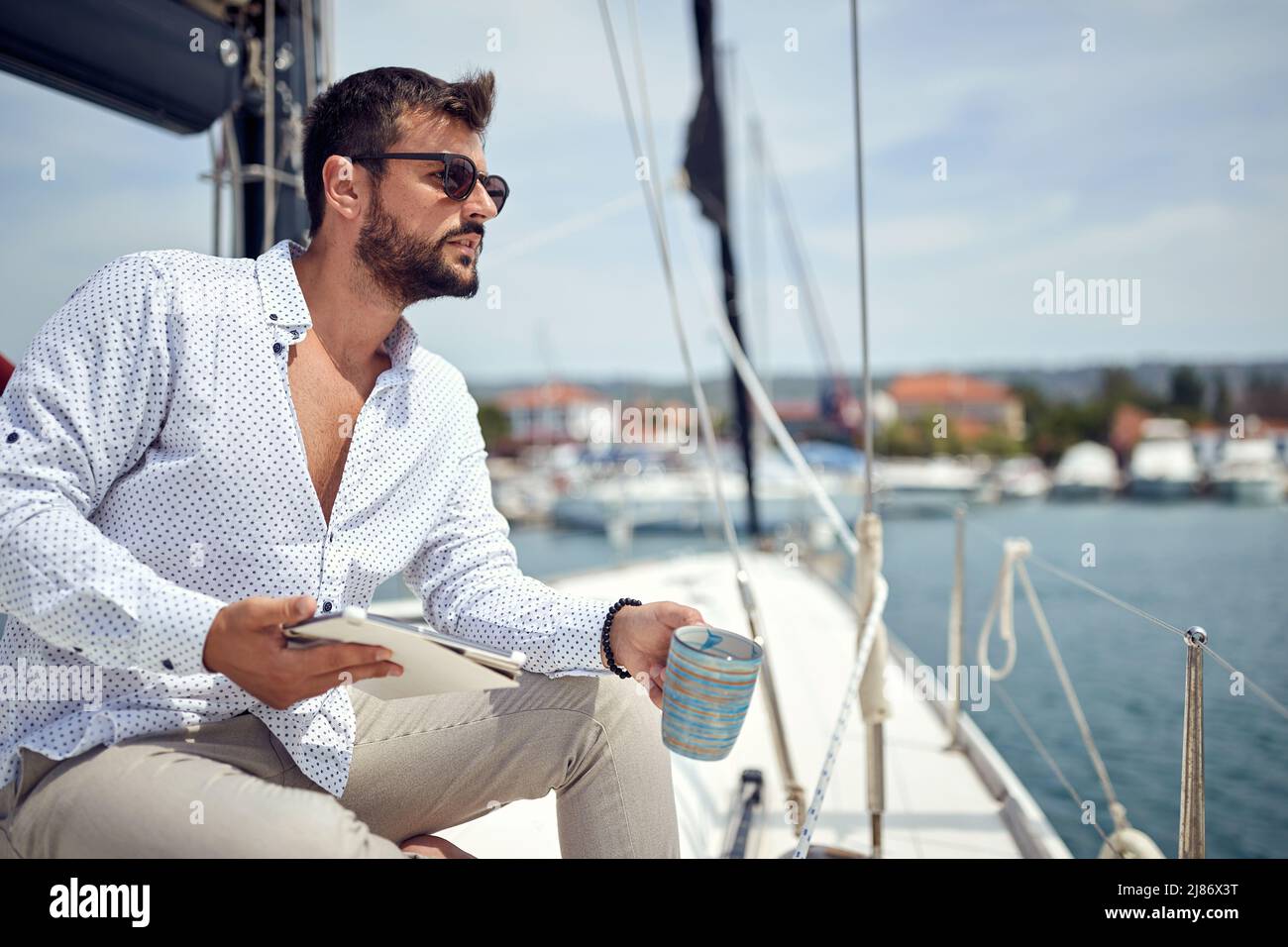 Un giovane uomo con un tablet è seduto e in posa per una foto sullo yacht mentre si gode un caffè e uno splendido scenario del molo in una giornata di sole su t. Foto Stock