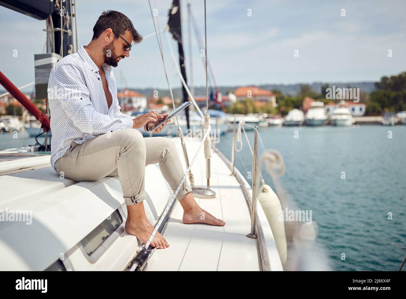 Un giovane uomo è seduto su uno yacht e utilizza un tablet mentre si guida attraverso il molo in una splendida giornata di sole sul mare. Estate, mare, vacanza Foto Stock