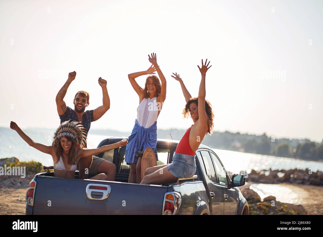 Un gruppo di giovani modelli belli seduti sul retro di un'auto e godersi la musica in una bella giornata di sole in mare. Estate, mare, vacanza, amicizia Foto Stock
