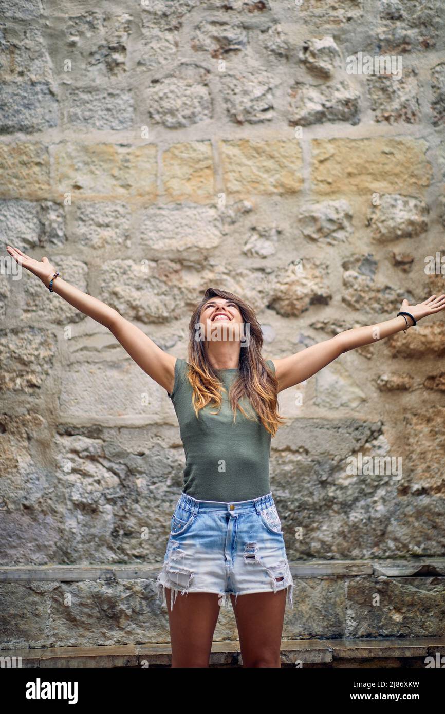 Giovane donna brunetta con braccia aperte in piedi e guardando in alto di fronte al muro di pietra. Felicità, libertà, concetto estivo. Foto Stock