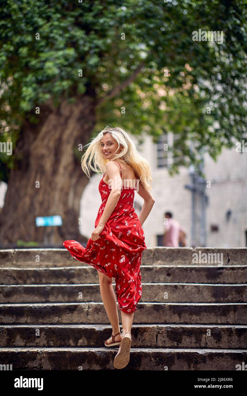 Una giovane ragazza in un abito rosso sta camminando i gradini nella città vecchia e posando per una foto in una bella giornata durante una vacanza sul mare. Vacanze, Foto Stock