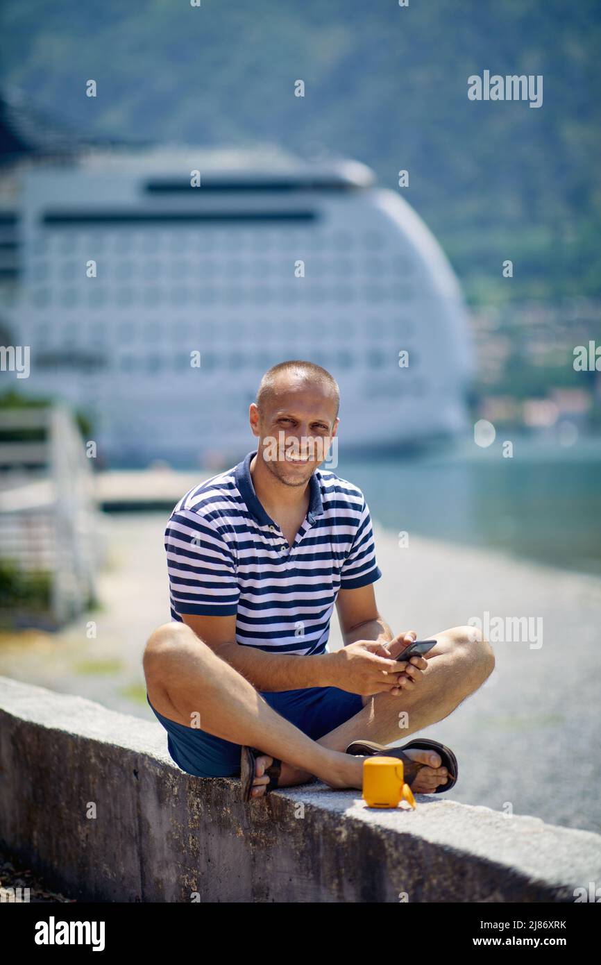 Uomo seduto sul ponte dall'acqua con smartphone, sorridente. Natura, Cruiser e montagne sfondo. Viaggi, affari, stile di vita Foto Stock