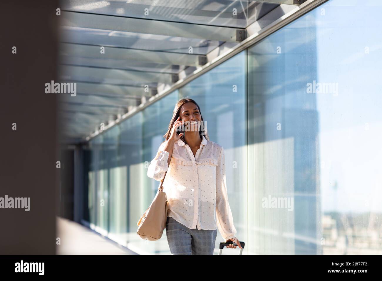 Sorridente donna d'affari biraciale che parla su smartphone camminando con i bagagli nel corridoio all'aeroporto Foto Stock