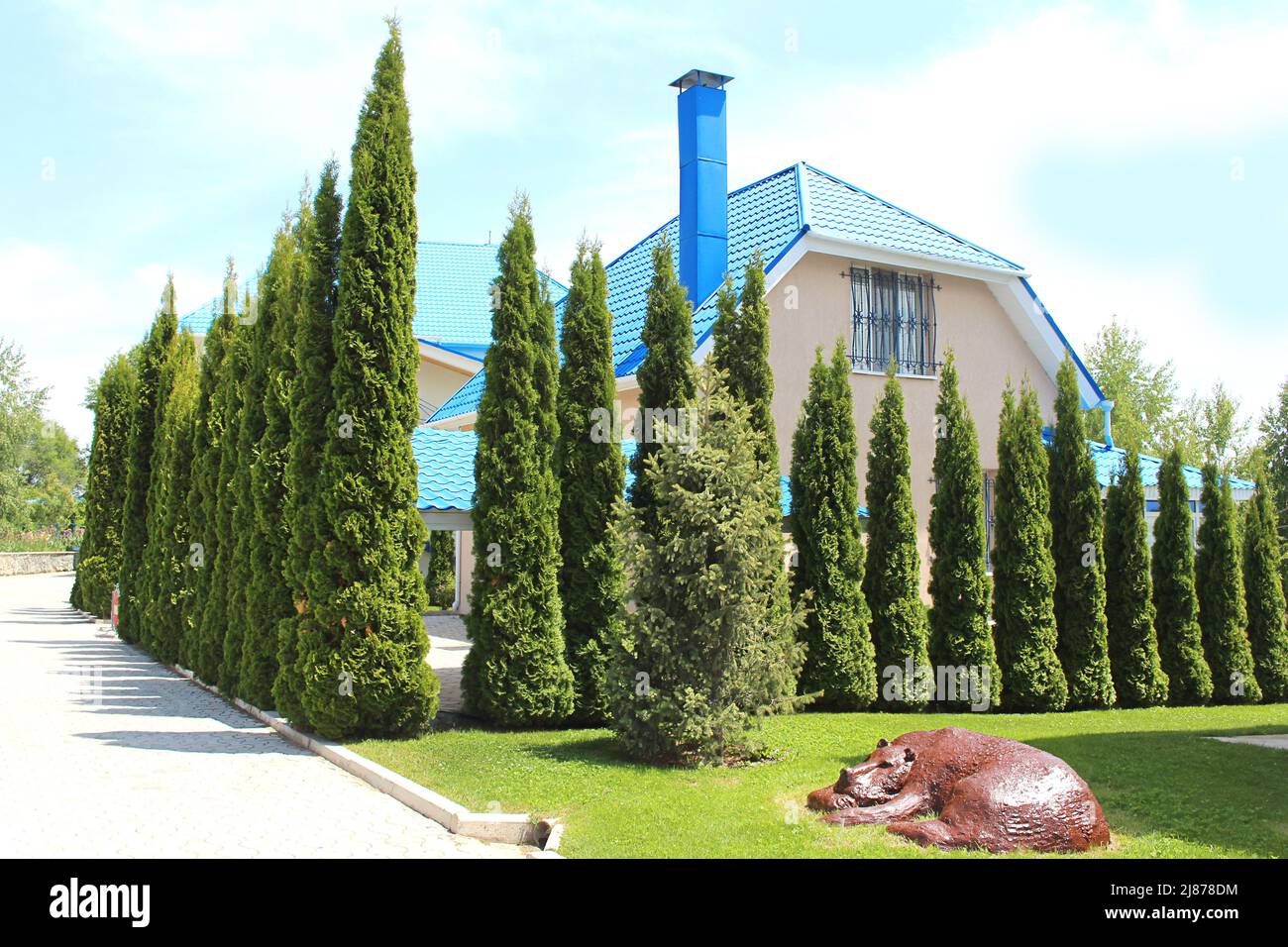 Casa con tetti blu con thujas verde e prato con statua di orso. Siepe in design paesaggistico Foto Stock