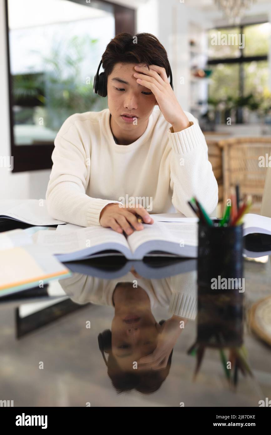 Ragazzo asiatico stressato adolescente con testa a mano indossando cuffie studiare sul tavolo a casa Foto Stock