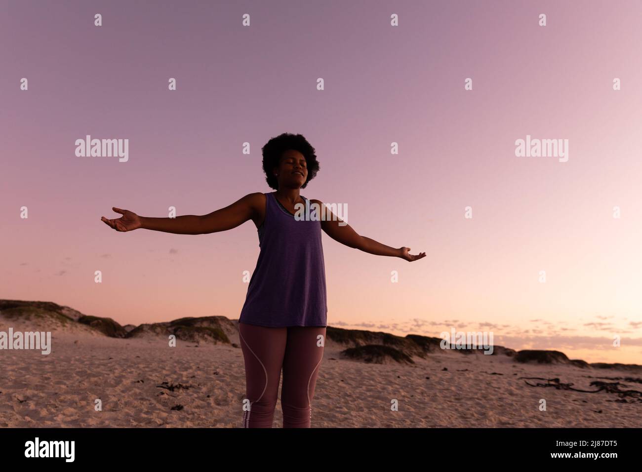 Donna matura afroamericana con capelli afro che chiudono gli occhi e allungano le braccia sulla spiaggia al tramonto Foto Stock