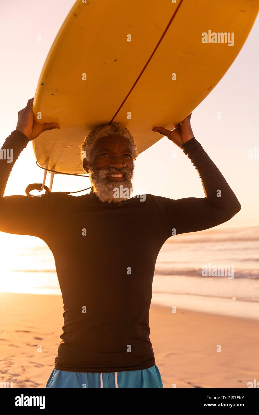 Sorridente uomo anziano afroamericano che porta la tavola da surf in testa contro il mare e il cielo limpido al tramonto Foto Stock