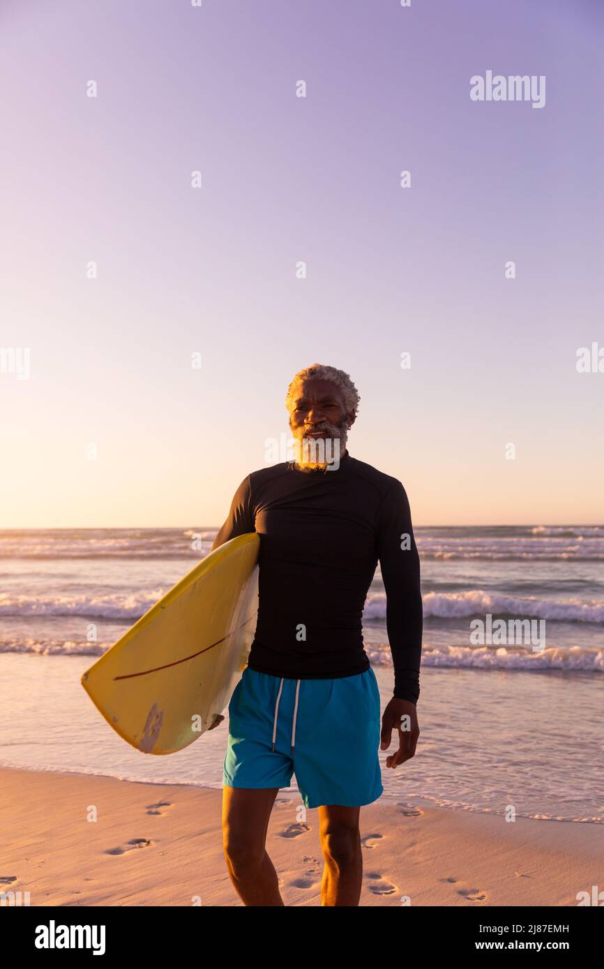 Uomo maggiore afro-americano con tavola da surf in piedi contro il mare e cielo limpido al tramonto Foto Stock