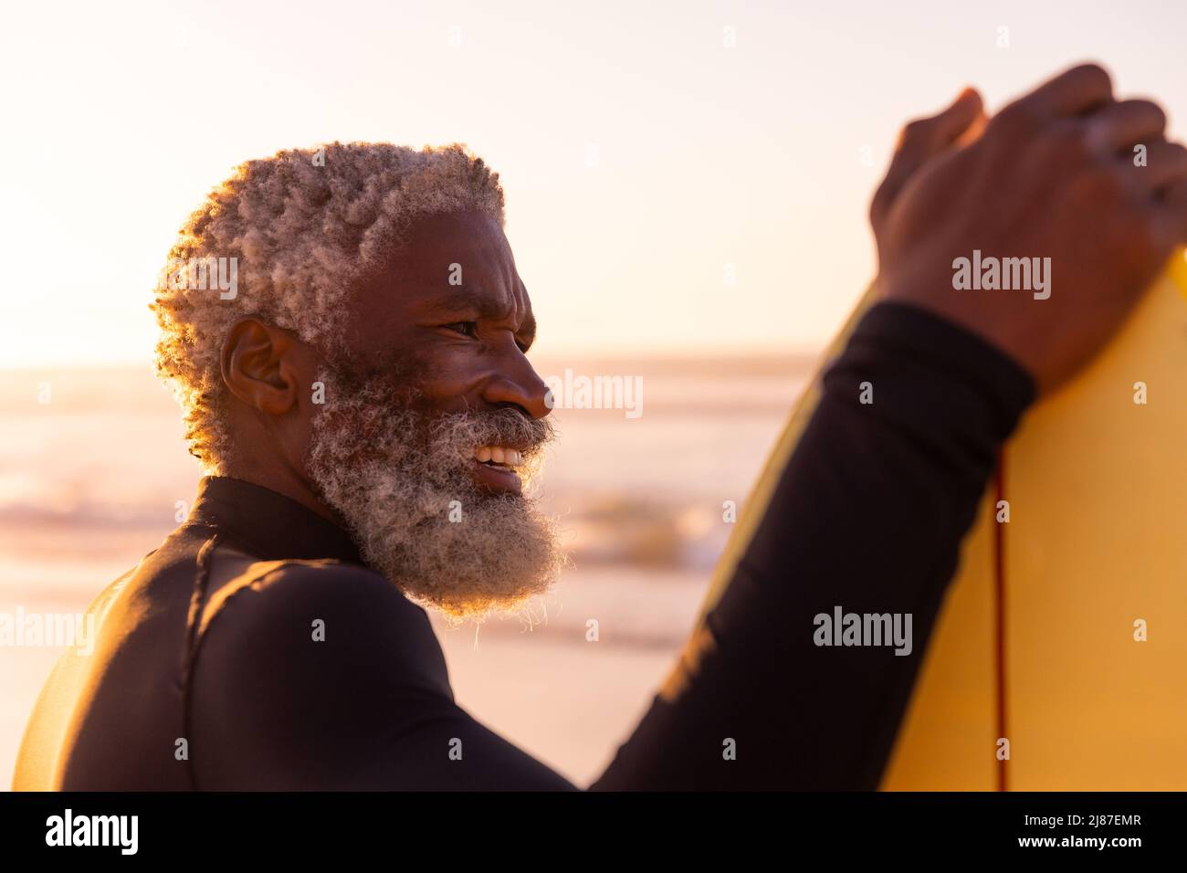 Sorridente uomo anziano afroamericano con tavola da surf che guarda via la spiaggia contro il cielo limpido Foto Stock