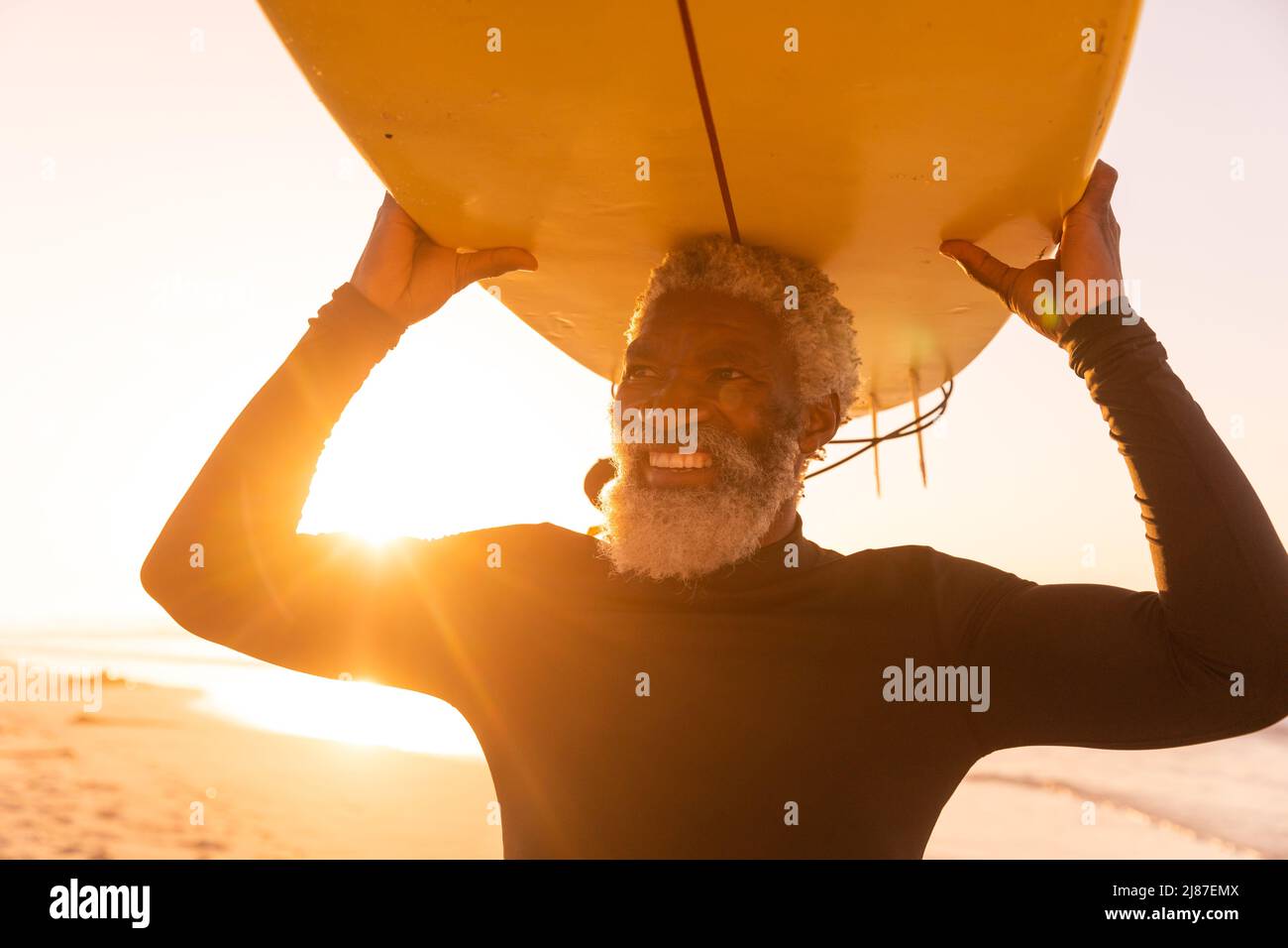 Sorridente uomo anziano afroamericano che porta la tavola da surf in spiaggia contro il cielo limpido al tramonto Foto Stock