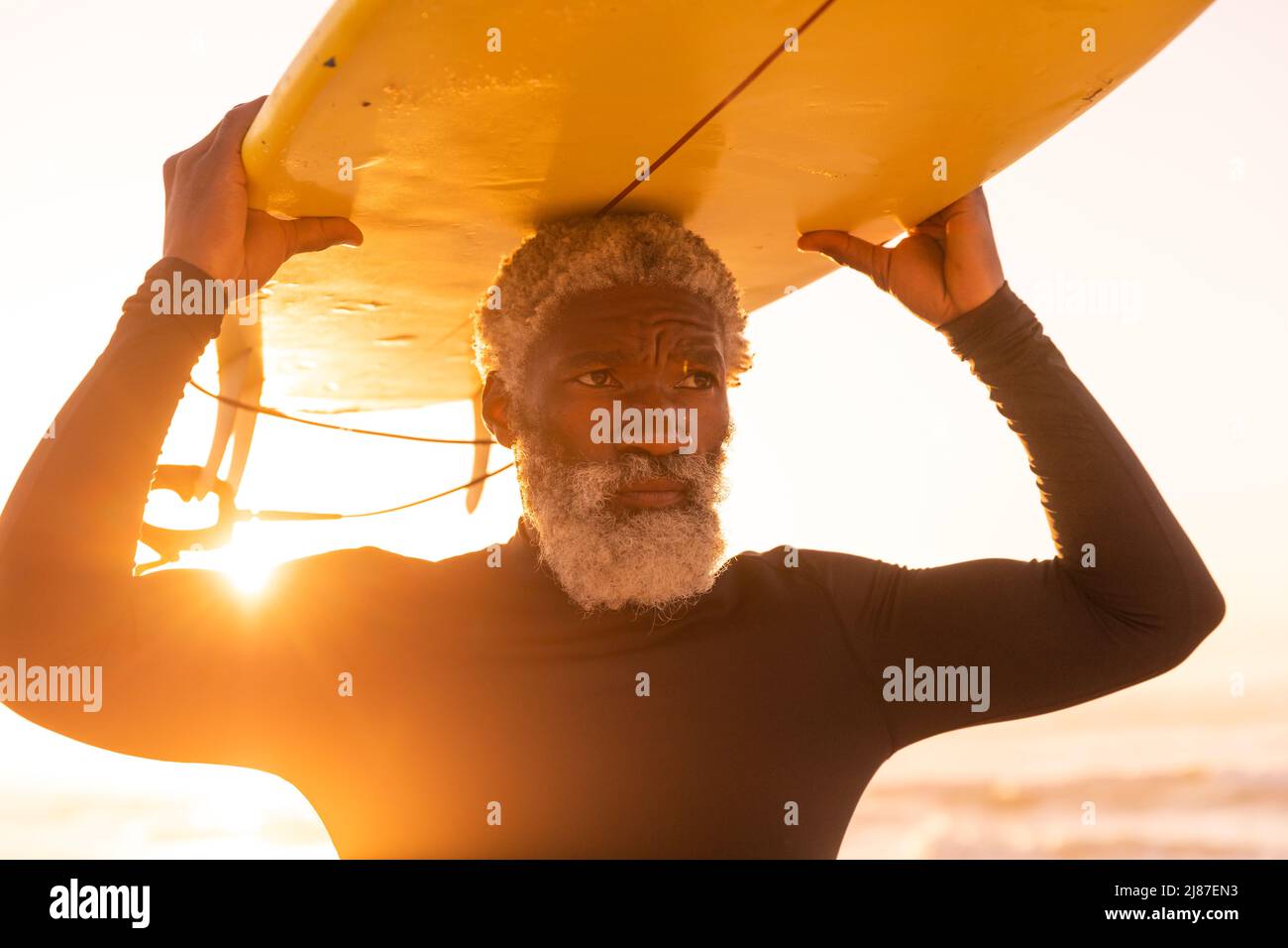 Uomo maggiore afroamericano portatore di surf in testa alla spiaggia contro cielo chiaro al tramonto Foto Stock