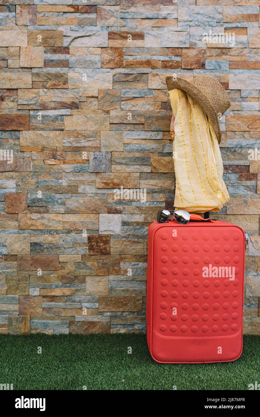 valigia davanti ad un muro di pietra e pavimento di erba per usare il testo. valigia con cappello, occhiali da sole e sciarpa. risorsa grafica. Foto Stock