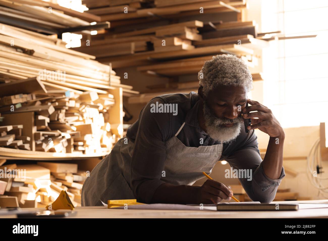 Falegname maturo afroamericano che parla su smartphone mentre disegnano il diagramma in officina Foto Stock