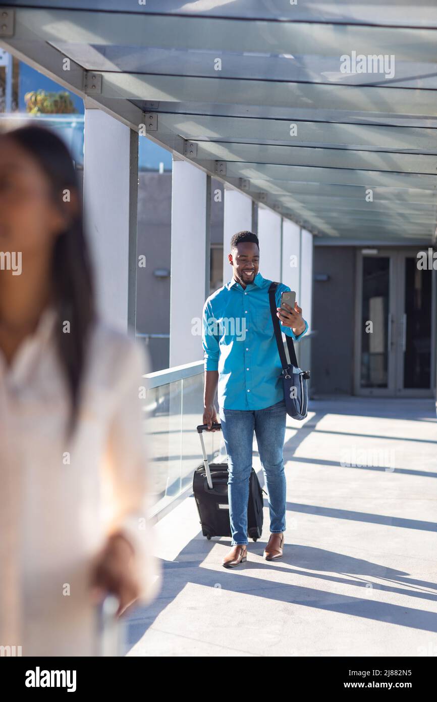 Sorridente uomo d'affari afroamericano che usa lo smartphone mentre cammina con i bagagli nel corridoio dell'aeroporto Foto Stock