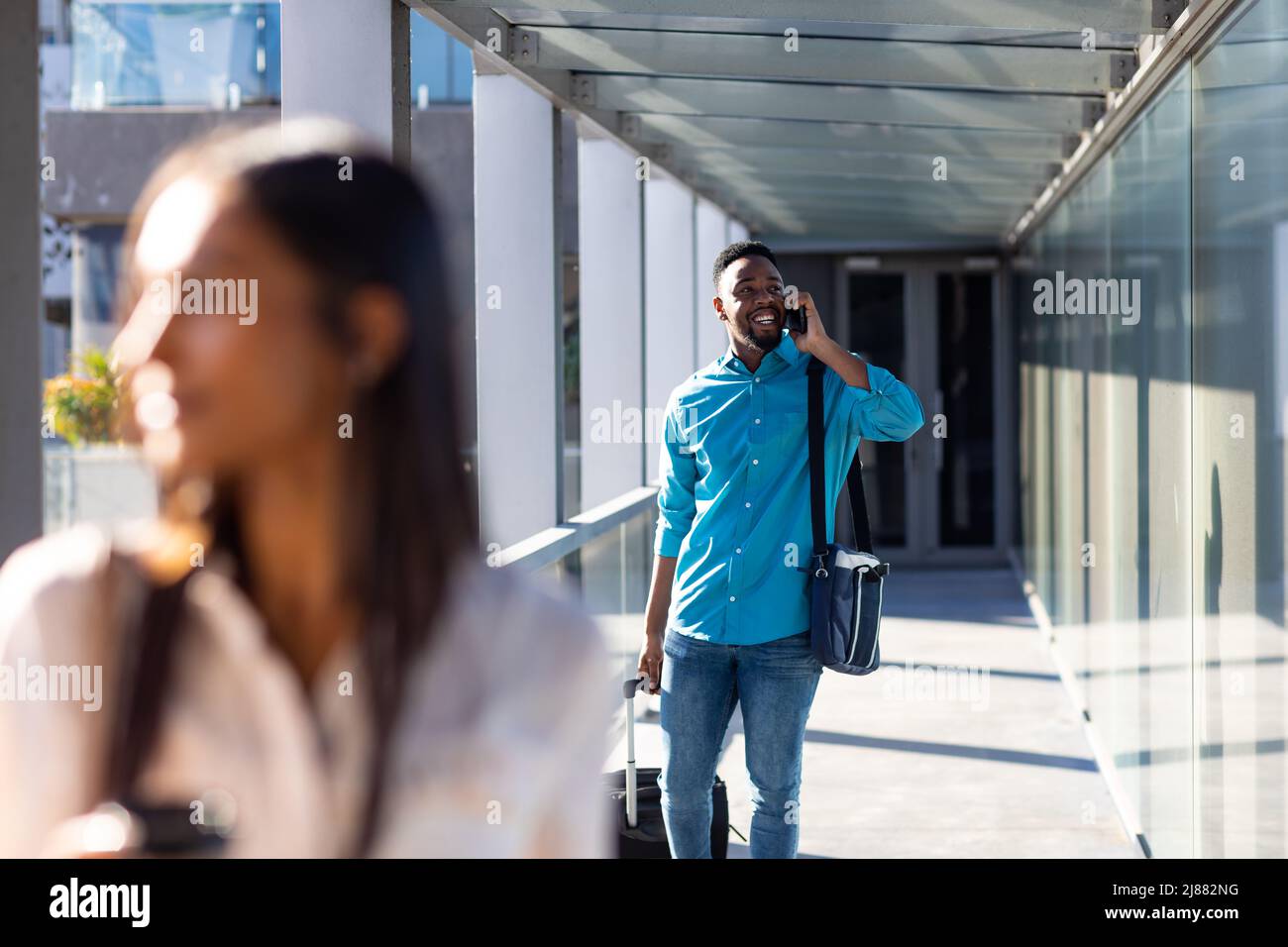 Un uomo d'affari afroamericano sorridente con i bagagli che parla al telefono cellulare nel corridoio dell'aeroporto Foto Stock