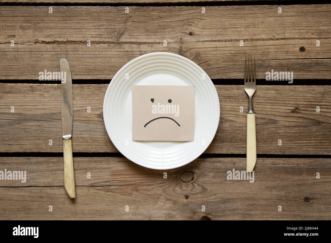 Una triste emoticon si trova in un piatto sul tavolo in cucina e accanto ad esso è un coltello con una forchetta, un cattivo umore, nessun cibo, la fame Foto Stock