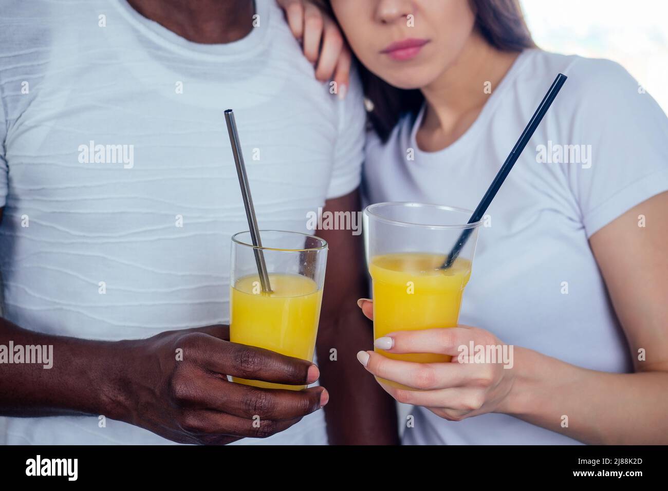 coppia internazionale in amore tenendo in mani occhiali con succo d'arancia.afro uomo utilizzare stoviglie monouso, ma donna europea using eco tubi d'acciaio Foto Stock