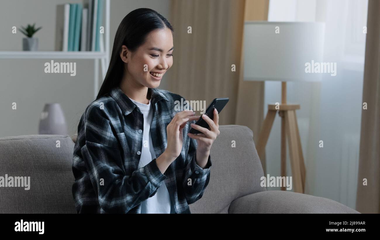 Asian felice sorridente ragazza donna seduta a casa divano guardando il telefono cellulare chattare navigazione utilizzando wi-fi web service guardare video online app SMS Foto Stock