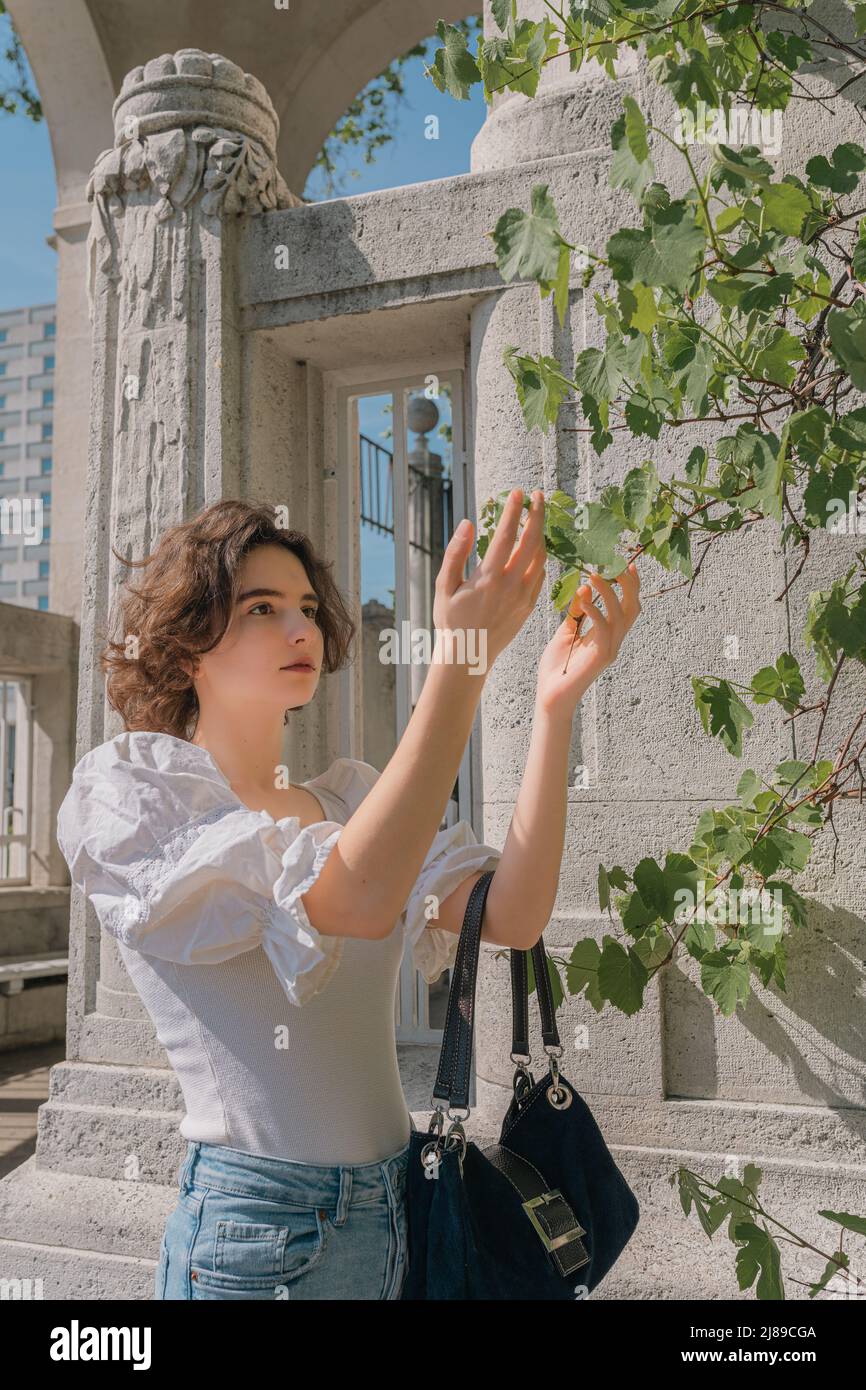 ragazza adolescente con capelli ricci marroni, figura snella, in abiti casual ed eleganti, stende belle mani a vite di uva che si snoda lungo il colum Foto Stock