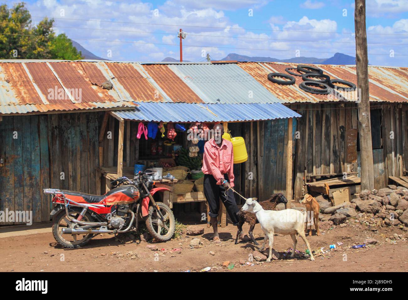 Bancarelle in legno con tetti di stagno arrugginito. Uomo con capre al negozio di stalla accanto alla moto. Pneumatici sul tetto. Village Street con pista di terra. Kenya, Africa Foto Stock