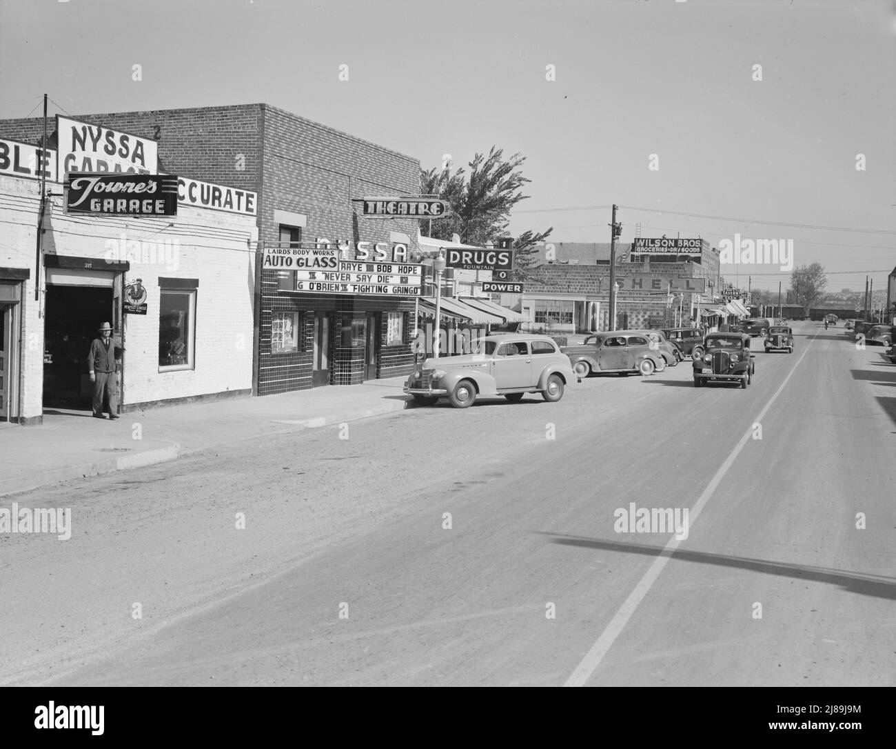 Strada principale di Nyssa, Oregon. Sabato pomeriggio. [Segni: 'Towne's Garage; Laird's Body Works - Auto Glass; Teatro [che mostra mai dire Die con Martha Raye e Bob Hope, e Fighting Gringo con George o'Brien); Drugs Fountain; Shell; Good Year; Wilson Bros Groceries - Dry Goods; Bob's Tavern'. Notare il treno merci all'attraversamento in piano]. Foto Stock