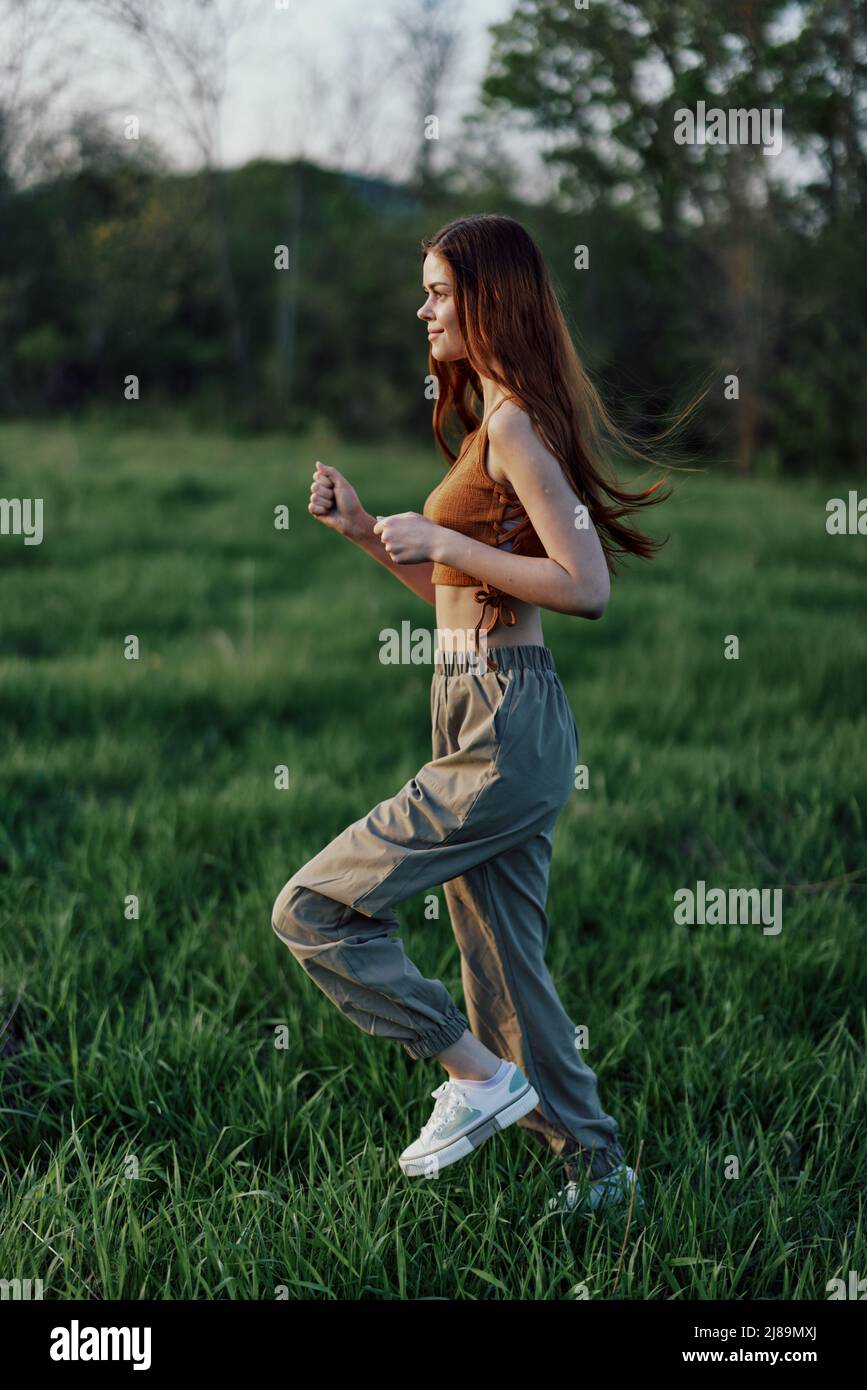 Una donna con lunghi capelli rossi lavora e corre sull'erba verde nel parco in pantaloni e sneakers Foto Stock