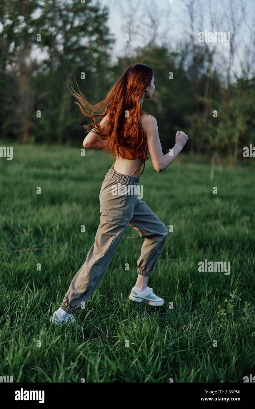 Una donna con lunghi capelli rossi lavora e corre sull'erba verde nel parco in pantaloni e sneakers Foto Stock