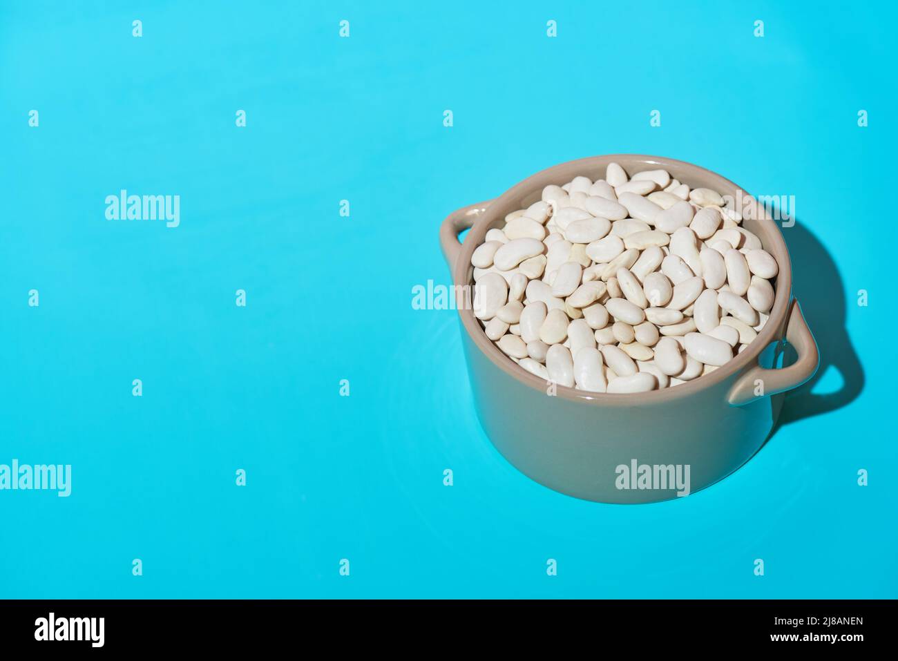 Fagioli bianchi secchi in un recipiente di vetro grigio su sfondo blu chiaro. Luce dura, ombra profonda. Primo piano, spazio di copia. Foto Stock